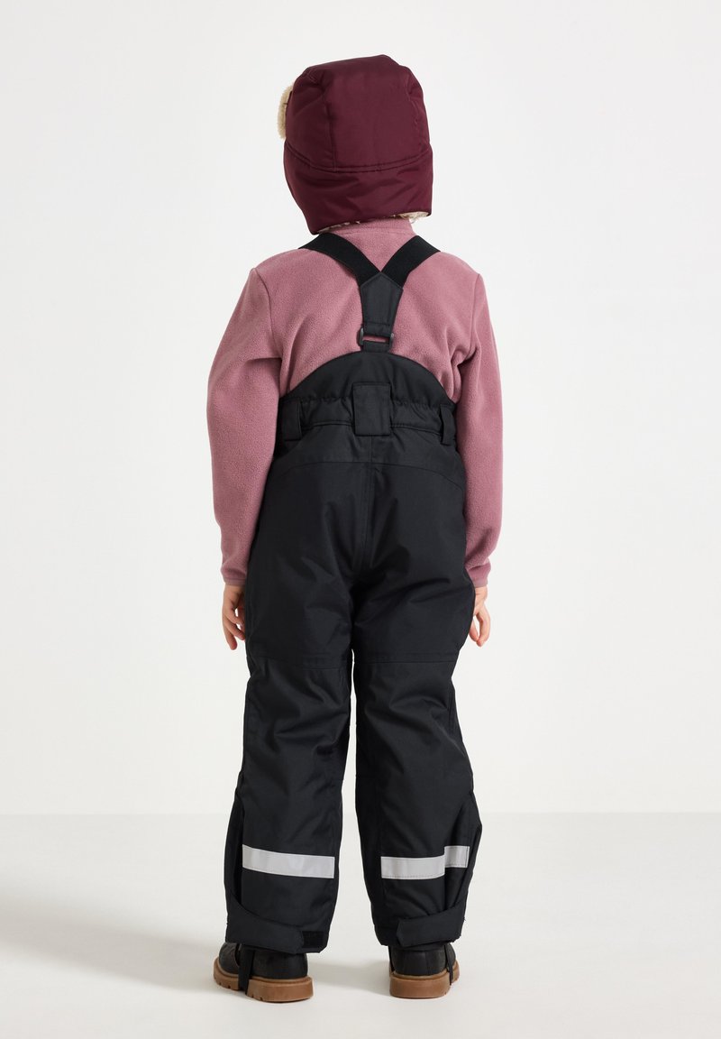 Sorte vandtætte overalls med reflekterende striber, iført over en lyserød fleece-top, suppleret af en kastanje farvet isoleret hat. Børns rygperspektiv.