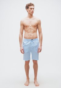 Νέος άνδρας χωρίς πουκάμισο, όρθιος ξυπόλητος, φοράει ανοιχτό μπλε και άσπρο ριγέ σορτς μέχρι το γόνατο, μπροστά σε λευκό απλό φόντο.