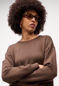 Brun sweatshirt med en afslappet pasform, ribbede manchetter og rund halsudskæring. Båret med ovale skildpaddesolbriller og gyldne hoopsøreringe.