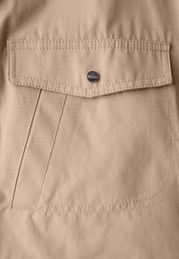 Béžová látka s texturovaným povrchem má klopu s kapsou a viditelným švem, uzavřenou kulatým kovovým patentem.