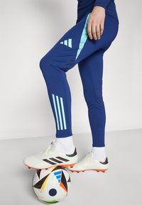 Leggings atletici blu con accenti azzurri e tre strisce verticali, abbinate a scarpini da calcio bianchi e neri, poggiati su un pallone da calcio a fantasia.