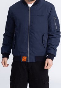 Homme portant un blouson bomber bleu marine avec fermeture éclair devant, poches en biais, poche zippée sur la manche et écusson orange au-dessus de la ceinture.