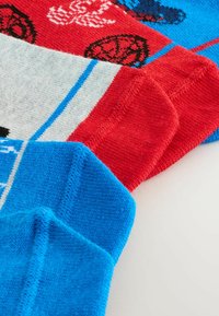 Gros plan sur trois chaussettes colorées avec des motifs sportifs bleus, rouges, blancs et à motifs, posées à plat sur une surface blanche.