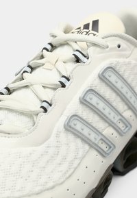 Close-up of beige Adidas sneaker with textured fabric, white laces with black stripes, three gray rubber stripes, and branded tongue.