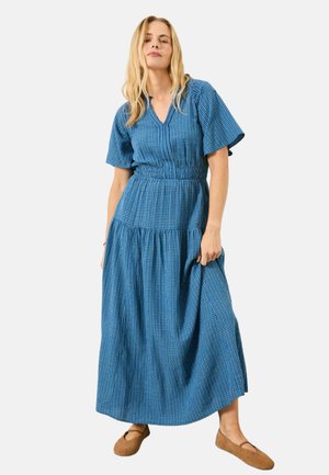 Frau trägt ein blau kariertes Maxikleid mit kurzen Ärmeln und beigefarbene flache Schuhe, steht vor einem einfarbigen Hintergrund.