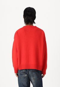 Pull rouge en tricot avec un ourlet et un col côtelés, présentant une coupe ample et un tissu texturé. Associé à un jean bleu.