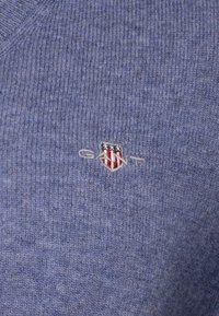 Blauer gestrickter Pullover mit V-Ausschnitt, gerippter Textur und kleinem gesticktem GANT-Logo mit einem Schild und Sternen auf der Brust.