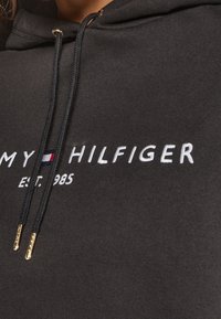 Haut à capuche noir avec le texte brodé blanc "TOMMY HILFIGER EST. 1985" accompagné d'un petit accent rouge et bleu. Tissu lisse avec des cordons de serrage.