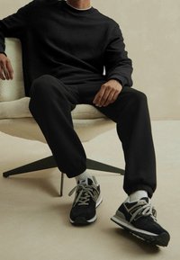 Persona seduta su una moderna poltrona beige, indossa una felpa nera a trama e pantaloni neri, calze bianche e sneaker New Balance nere e grigie.