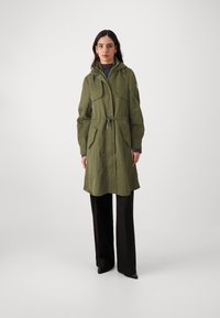 Parka verde oliva con cappuccio, vita aderente, due ampie tasche frontali e vestibilità comoda. Tessuto testurizzato con finitura opaca.