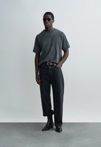 Mees, kes kannab tumedat halli T-särki, musti teksaseid vöökohaga, musti saapaid ja ümmargusi päikeseprille, seisab helehallil ühevärvilisel taustal.