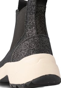 Botín negro texturizado hasta el tobillo con costura elástica a los lados, detalle trenzado en la parte trasera y suela de goma de color crema. Presenta un diseño robusto y moderno.