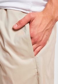 Pantaloni bej deschis cu o textură netedă, având un bochet lateral. O mână este plasată în interiorul bochetului, evidențiind căderea țesăturii.