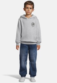 Grå huvtröja med framficka, prydd med en rund svart logotyp. Bärs tillsammans med lösa blå denimbyxor och vita sneakers.