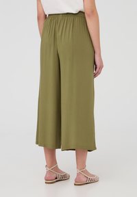 Pantaloni a gamba larga verde oliva con vita elastica, texture liscia e vestibilità drappeggiata. Abbinati a sandali beige con cinturini.