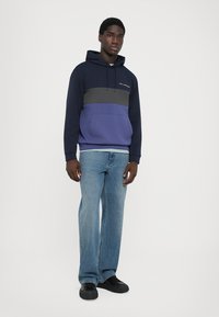 O sweatshirt com capuz apresenta um design em blocos de cor nas cores azul escuro, cinza e roxo, com um bolso canguru e capuz ajustável. Combinado com jeans azul claro.