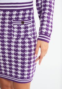 Paarse en witte gebreide jurk met een houndstooth patroon. Heeft een zak met een gouden knoop en gestreepte manchetten bij de halslijn en de zoom.