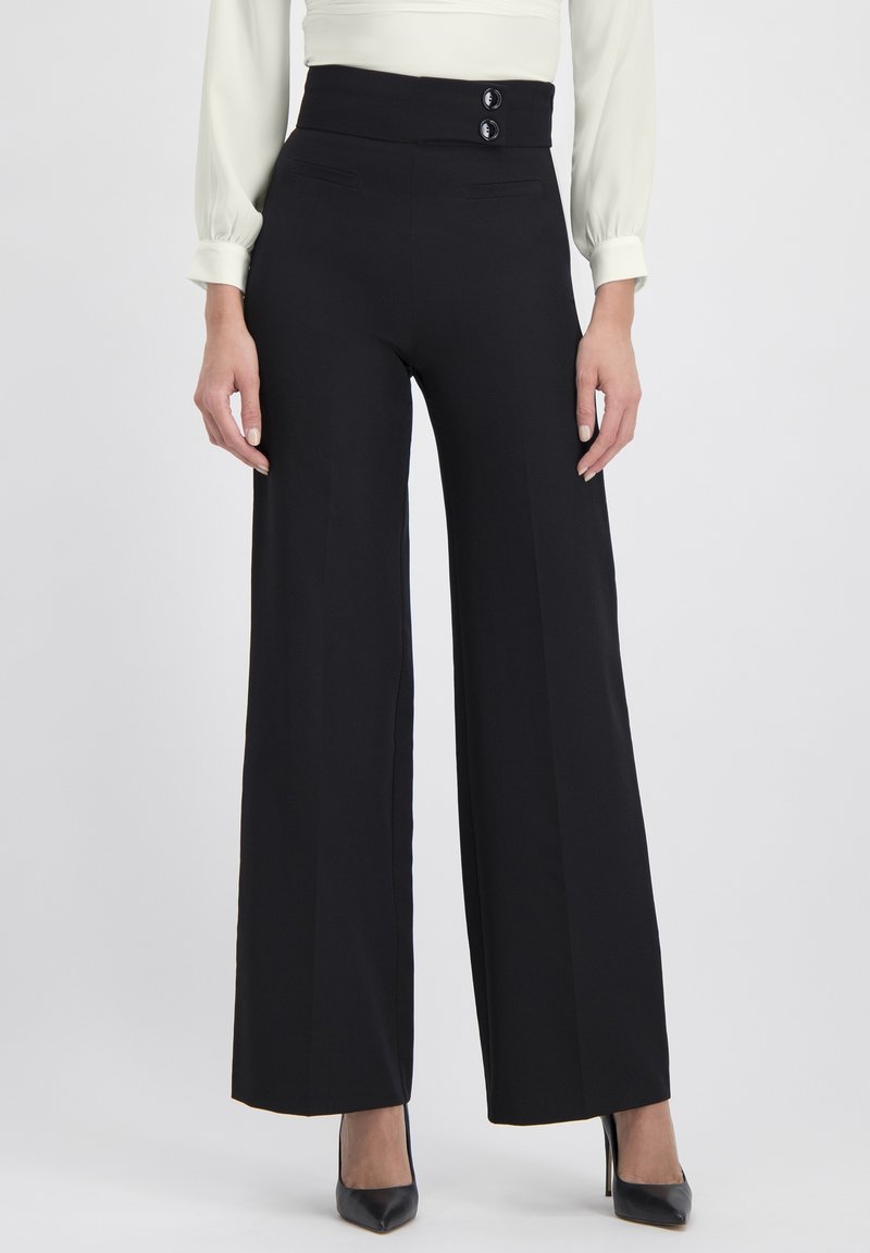 Mujer que lleva pantalones negros de pierna ancha de talle alto con dos botones en la cintura y tacones negros de punta afilada.