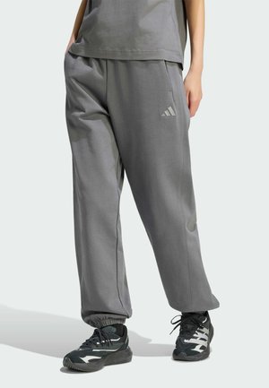 Osoba nosząca luźne, szare spodnie dresowe Adidas z kieszeniami, w połączeniu z czarno-białymi sportowymi butami, stojąca na jasnym tle.