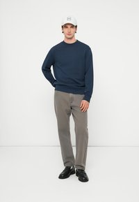 Tamsiai mėlynas megztinis su ilgomis rankovėmis, derinamas su šviesiai pilkomis kelnėmis. Modelis dėvi baltą kepurę su logotipu ir juodus batus.