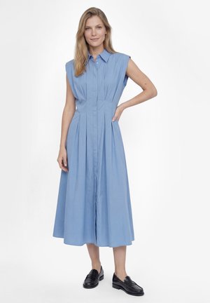 Kobieta stojąca z ręką na biodrze, ubrana w niebieską, bezrękawną, midi sukienkę i czarne mokasyny na białym tle.