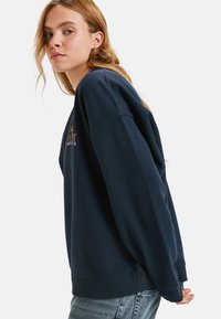 Granatowy sweter z ściągaczami na rękawach i dole, z kolorowym haftowanym tekstem na przodzie. Lekko oversize'owy krój o miękkiej fakturze.