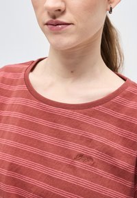 Κοντινό πλάνο ενός ατόμου που φοράει κόκκινο πουκάμισο με ρίγες, στρογγυλή λαιμόκοψη και μικρό κεντημένο λογότυπο στο στήθος, καθώς και ασημένιο σκουλαρίκι κρίκο.