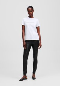 Weißes Baumwoll-T-Shirt mit dem Text "KARL LAGERFELD", kombiniert mit schwarzen, taillierten Hose aus Kunstleder und schwarzen Slippers mit Verzierungen.