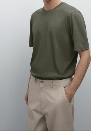 Homme portant une chemise à manches courtes vert olive rentrée dans un pantalon beige, une main dans la poche, sur un fond uni.