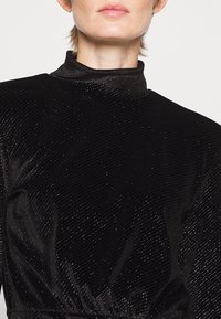 Robe noire à col montant en tissu texturé avec des accents métalliques. Elle présente une taille cintrée et un éclat subtil avec une surface scintillante.
