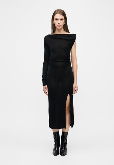 Femme debout portant une robe noire asymétrique avec une manche longue et une fente haute sur la cuisse, assortie à des bottines noires, sur un fond blanc.