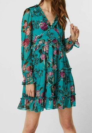 Türkisfarbenes, langärmliges Blumenkleid mit Rüschen, V-Ausschnitt und gestuftem Rock, getragen von einer Frau mit offenem Haar.