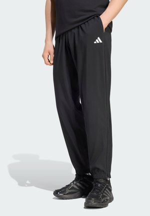 Osoba nosząca czarne spodnie sportowe Adidas z elastycznymi ściągaczami i czarne buty sportowe, stojąca z jedną ręką w kieszeni.