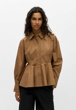 Femme aux longs cheveux bouclés portant une chemise marron boutonnée avec une taille évasée et un pantalon noir, debout devant un fond uni.