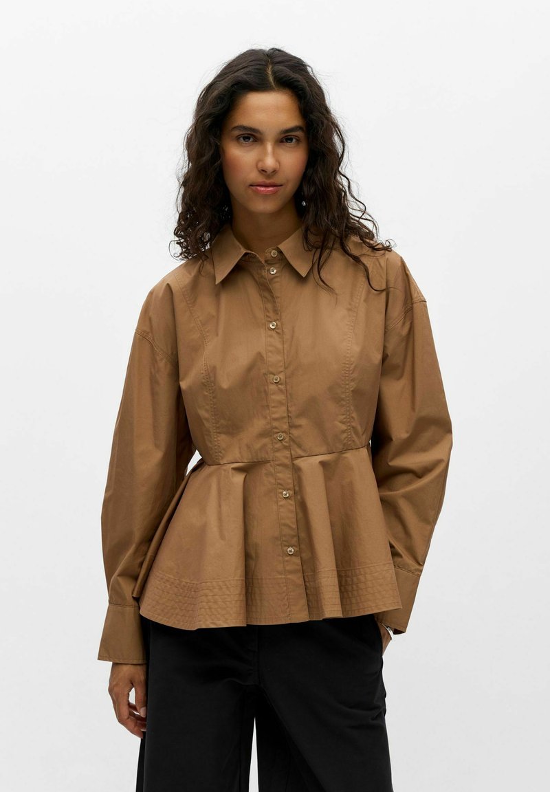 Femme aux longs cheveux bouclés portant une chemise marron boutonnée avec une taille évasée et un pantalon noir, debout devant un fond uni.