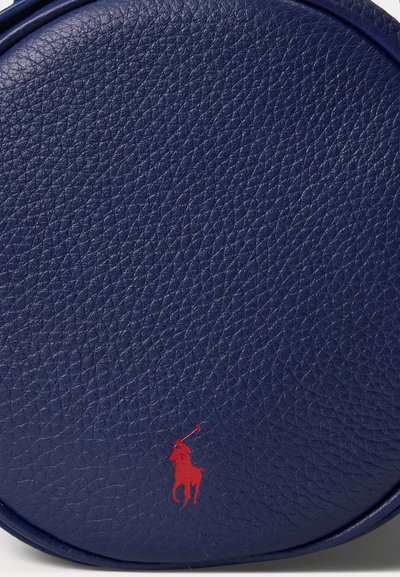 Tumši zila ādas apaļa soma ar teksturētu virsmu un sarkanu logotipa detaļu. Aprīkota ar gludu rāvējslēdzēju un elegantu, neuzkrītošu dizainu.