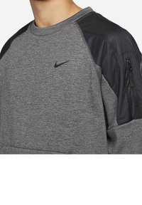 Homme portant un sweat-shirt Nike gris avec des panneaux noirs aux épaules et une poche zippée sur la manche gauche, gros plan sur le haut du torse.