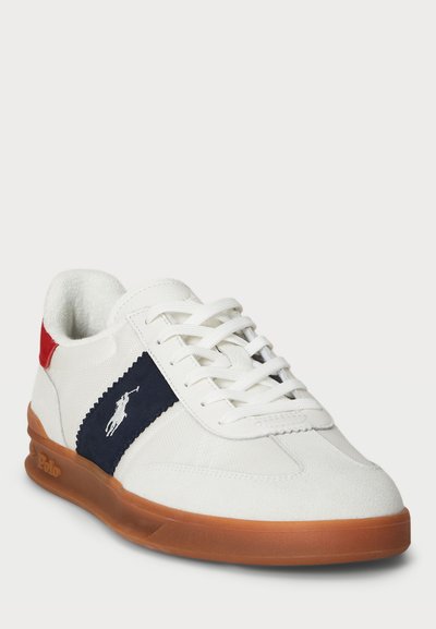 Sapatilhas baixas brancas com um cabedal em camurça e lona, painel lateral azul marinho, detalhe no calcanhar em vermelho e sola de borracha castanha. Apresenta atacadores planos.