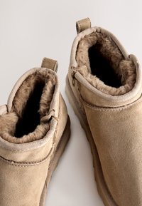 Bottines en daim beige avec doublure en fausse fourrure, fermetures éclair latérales et languettes de talon renforcées, présentant une texture douce et un design robuste.