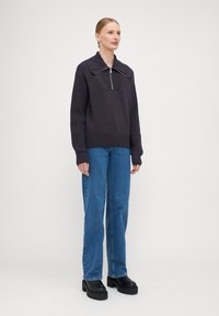 Mørk grå strikkegenser med glidelås og høy krage, sammen med blå denimjeans og tykke svarte platå-sko. Flat tekstur, avslappet passform.