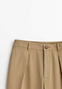 Pantalons beiges avec fermeture à bouton, dotés de plis à la taille, fabriqués à partir d'un tissu texturé. Le design comprend des passants de ceinture et des détails cousus.