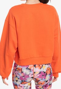 Sudadera corta naranja con cuello redondo y hombros caídos, combinada con leggins florales multicolores que presentan una textura translúcida.