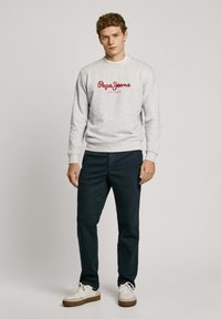 Sudadera gris con logo rojo "Pepe Jeans", combinada con pantalones navales y zapatillas blancas. Tejido suave, diseño casual, puños y dobladillo acanalados.