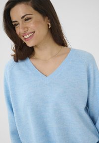 Femme souriante avec des cheveux bruns portant des boucles d'oreilles en or et un délicat collier en chaîne dans un pull V bleu clair sur fond blanc.