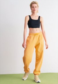 Top corto negro y pantalones de chándal amarillos con cinturilla y puños elásticos. El modelo lleva zapatillas neutras. Fondo liso.