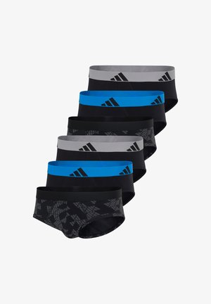 Atletyczny mężczyzna stojący boso, ubrany w czarne slipy z niebieskim pasem w talii, na którym znajduje się czarne logo Adidas, patrzący lekko w lewo.