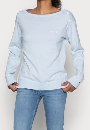 Femme portant un pull bleu clair à manches longues avec des boutons sur les épaules et un jean bleu clair, debout avec les mains près des poches.