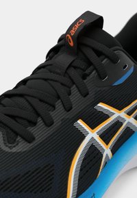 Zapatilla deportiva negra con parte superior texturizada, con marca naranja y acento azul. Presenta líneas de diseño en blanco y amarillo para un detalle adicional.