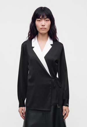Mulher com cabelos pretos e longos veste uma blusa preta tipo envelope com gola de pala branca e saia de couro preta contra um fundo claro e simples.
