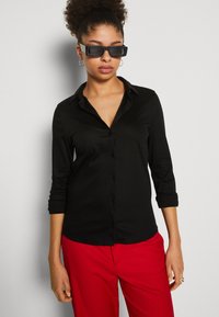 Camicia nera a maniche lunghe con bottoni in tessuto liscio, caratterizzata da un colletto classico e polsini arrotolati, abbinata a pantaloni rossi vivaci.